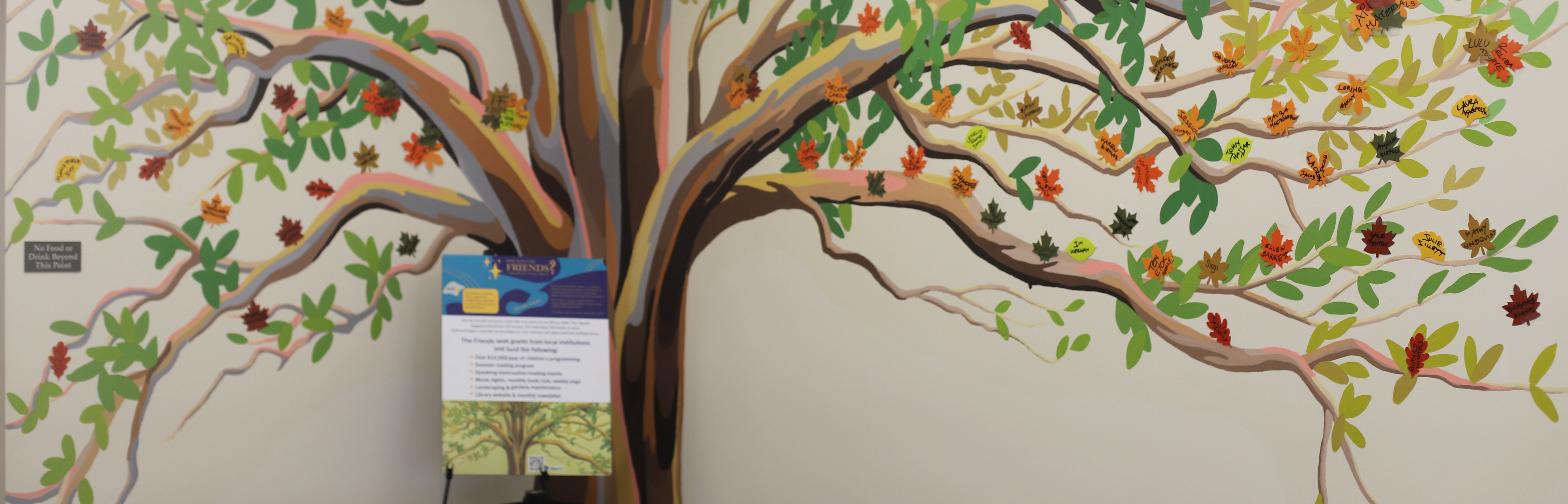 Photo of the Friends' tree mural in the lobby of the library with a sign explaining the Friends' mission