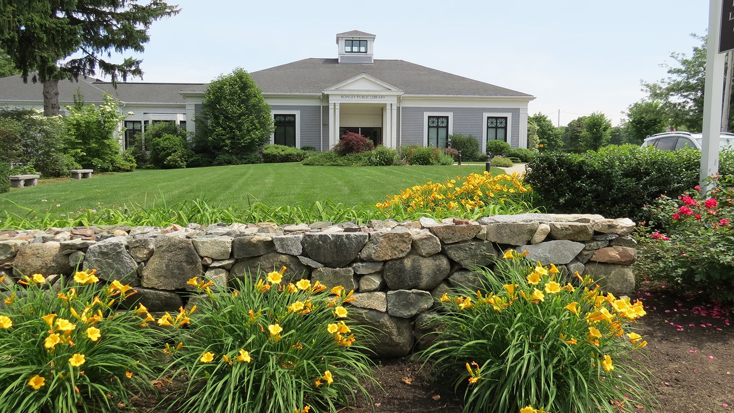 Photo of the front exterior of the Rowley Public Library including the garden and flowers.